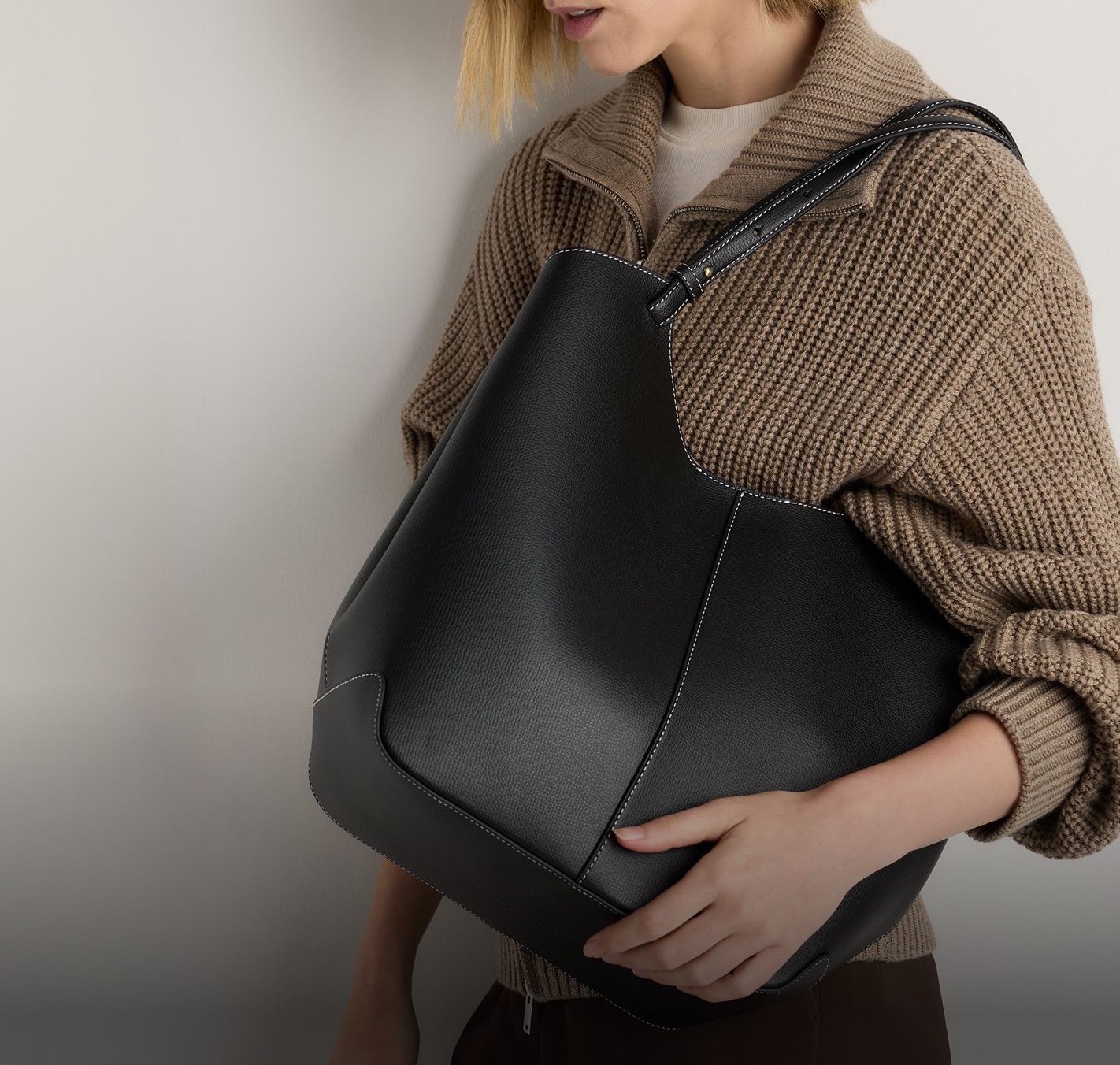Model in a brown ribbed knit cardigan carrying a black leather Rowan tote with white stitching — Container 3 banner, UK homepage, 22 Oct 2026