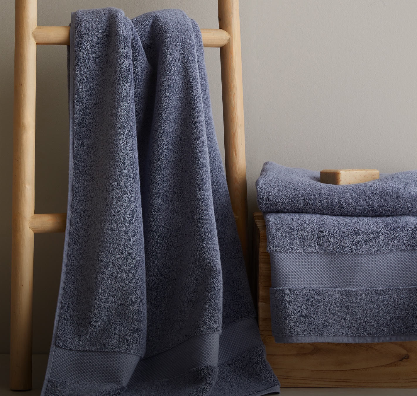 Mineral blue-grey Turkish Ultra Plush towels draped over a wooden ladder with a stack of folded towels and a bar of soap — Container 2 banner, UK homepage, 15 Oct 2026